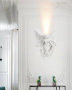 Fairy-shaped Clérachic wall light lit on a tall white wall with ornate plasterwork and two small green figurines on a console.