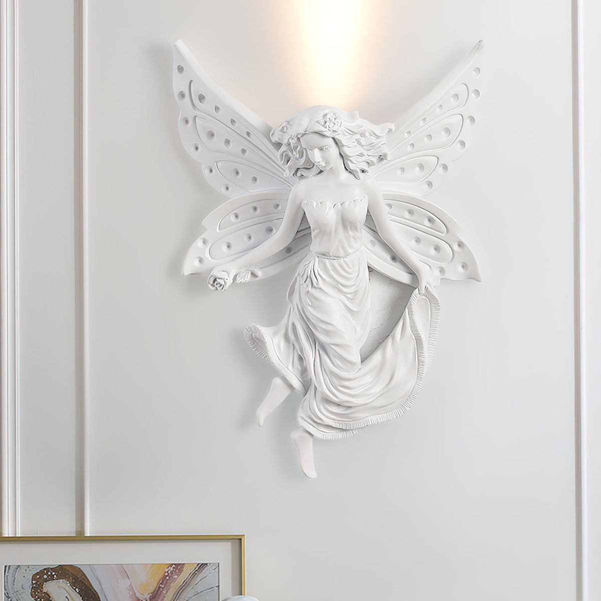 Large view of a lit fairy-shaped Clérachic wall light on a white wall, with the edge of a gold-framed picture at the bottom.