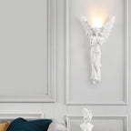 Standing-angel Clérachic wall light lit on a white panelled wall above a white bust and a teal cushion.