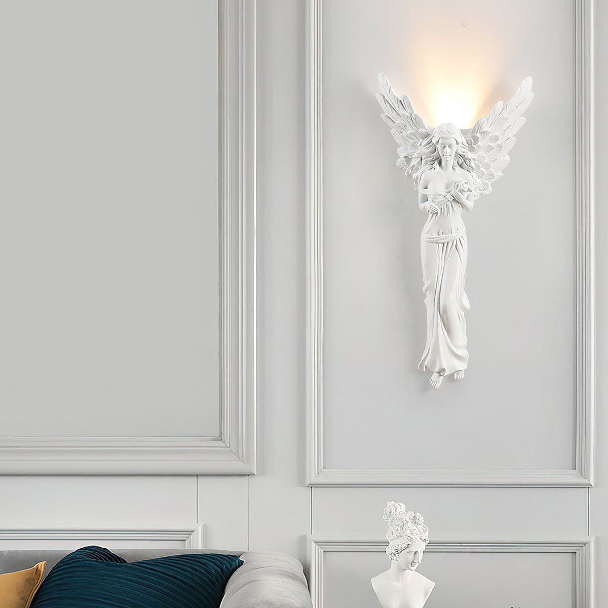 Standing-angel Clérachic wall light lit on a white panelled wall above a white bust and a teal cushion.