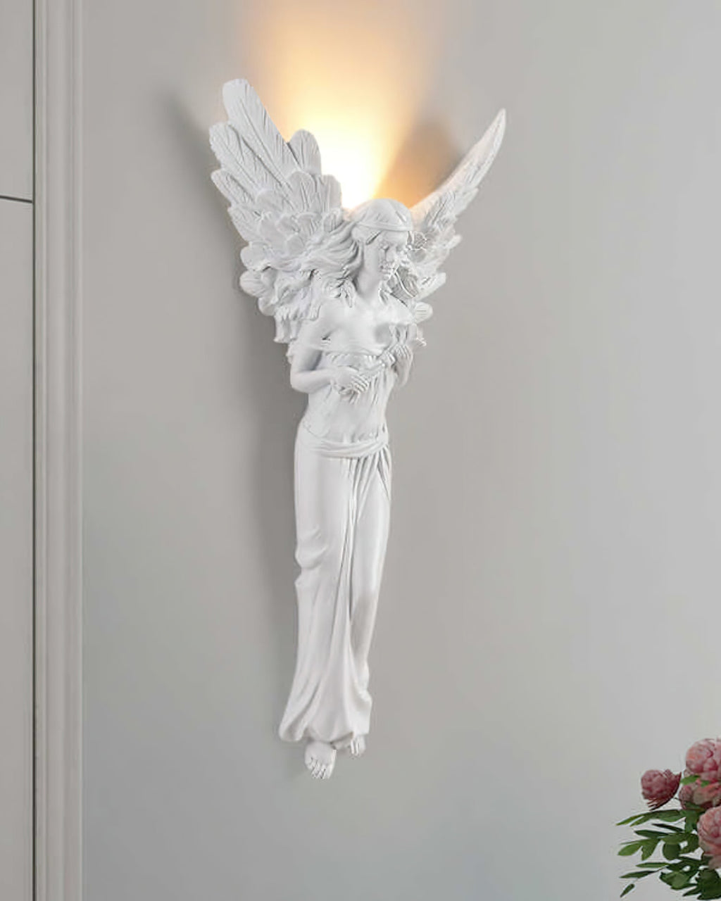 Standing-angel Clérachic wall light lit on a grey wall, with pink flowers in the lower right corner.