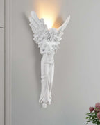 Standing-angel Clérachic wall light lit on a grey wall, with pink flowers in the lower right corner.
