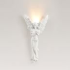 Standing-angel Clérachic wall light lit on a pale beige wall, a warm glow rising above its feathered wings.