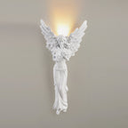 Lit standing-angel Clérachic wall light on a warm grey wall, the glow from above its wings softly lighting the wall.