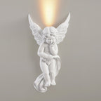 Cherub Clérachic wall light, lit, on a grey wall, a bright warm glow above its feathered wings.