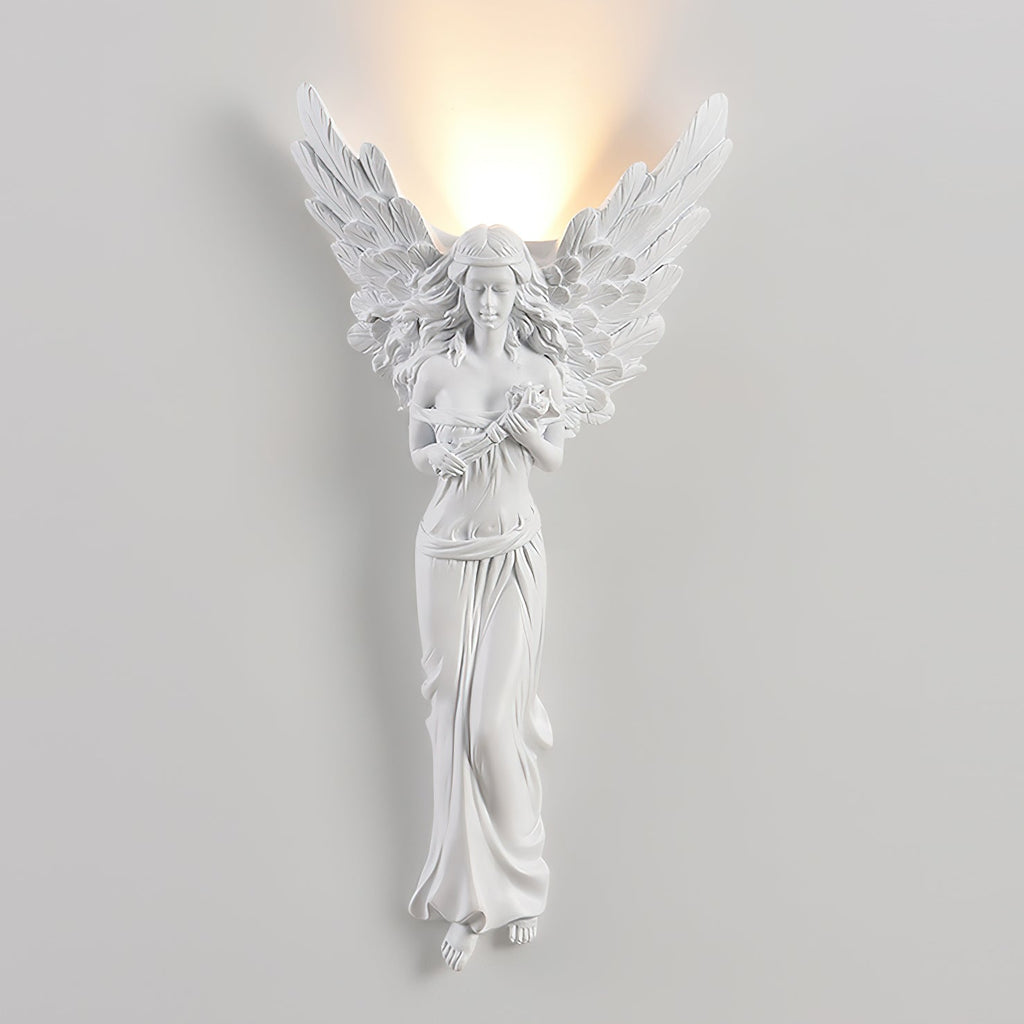 Lit standing-angel Clérachic wall light on a pale wall, with warm light washing up the wall from behind its wings.