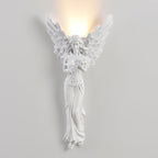 Lit standing-angel Clérachic wall light on a pale wall, with warm light washing up the wall from behind its wings.