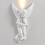 Lit cherub Clérachic wall light on a pale wall, with warm light rising from behind its feathered wings.