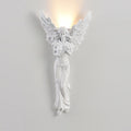 Standing-angel Clérachic wall light, lit, on a pale wall, a soft warm glow rising above its feathered wings.