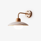 Clérachic wall light with a travertine dome shade, a curved brass-toned arm and a round wooden wall plate on a plain backdrop.