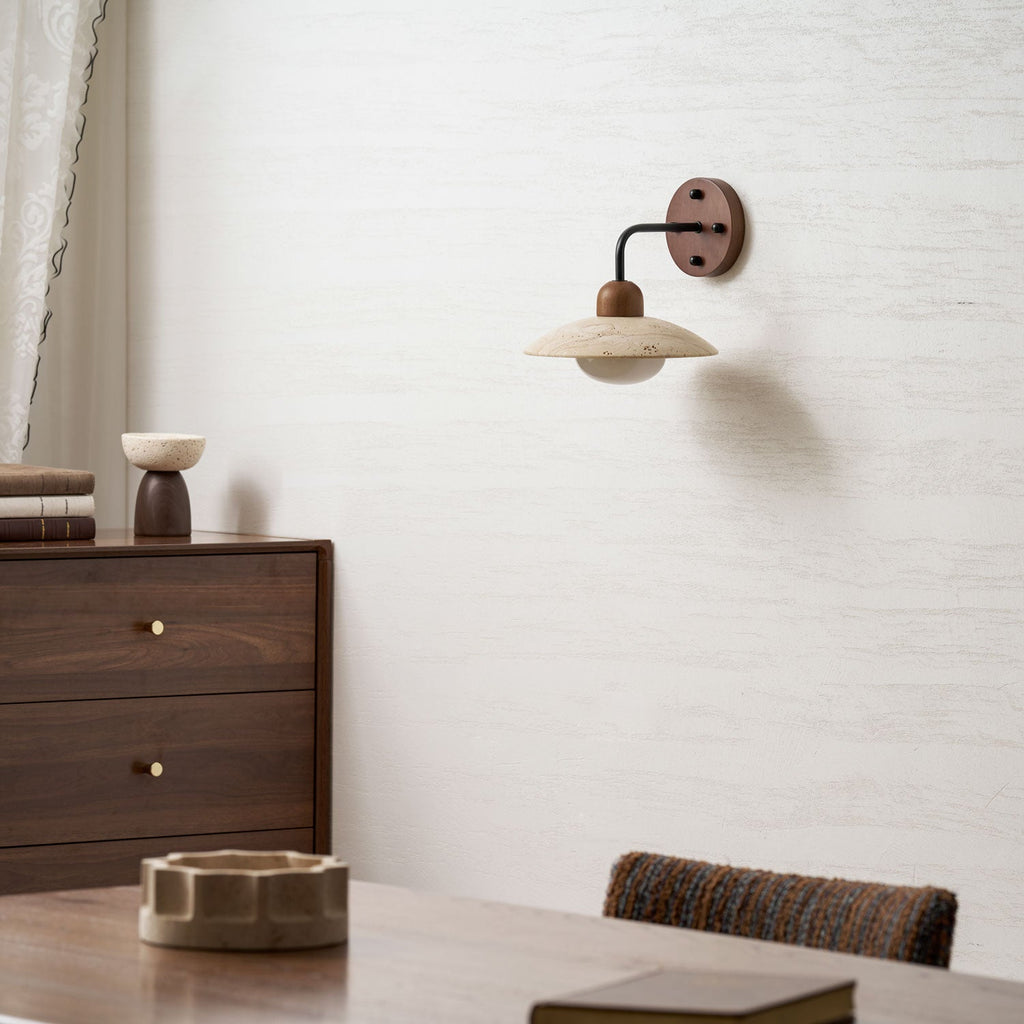 A small Clérachic wall light with a travertine shade on a textured white wall above a walnut dresser and dining table.