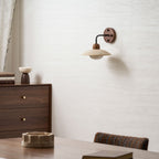 A small Clérachic wall light with a travertine shade on a textured white wall above a walnut dresser and dining table.