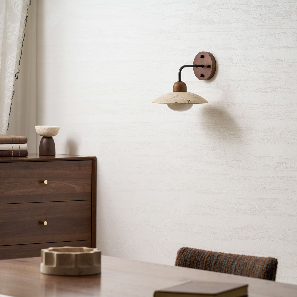 A small Clérachic wall light with a travertine shade on a textured white wall above a walnut dresser and dining table.