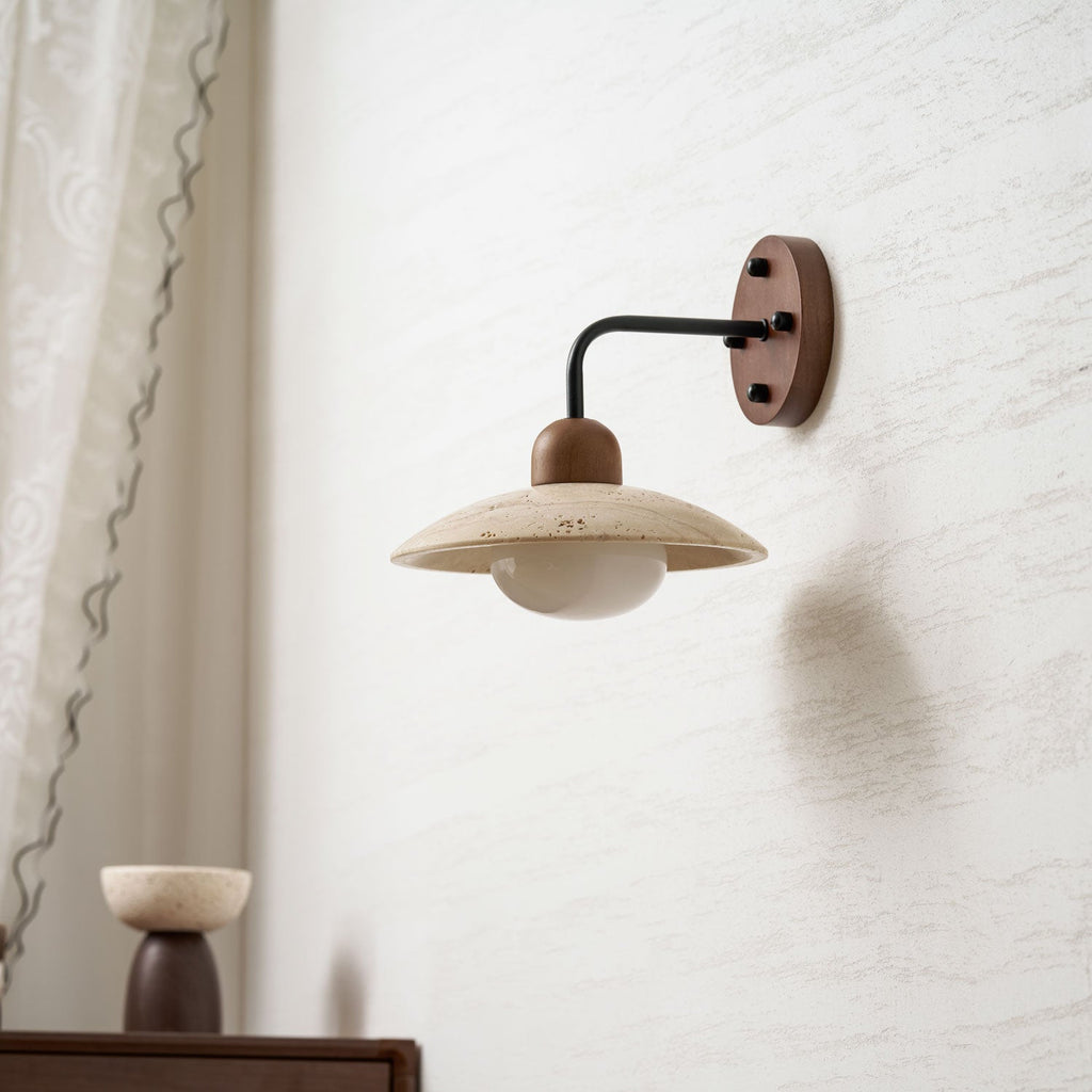 Clérachic wall light with a black arm and a wooden wall plate, its travertine dome unlit against a textured pale wall.