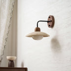 Clérachic wall light with a black arm and a wooden wall plate, its travertine dome unlit against a textured pale wall.
