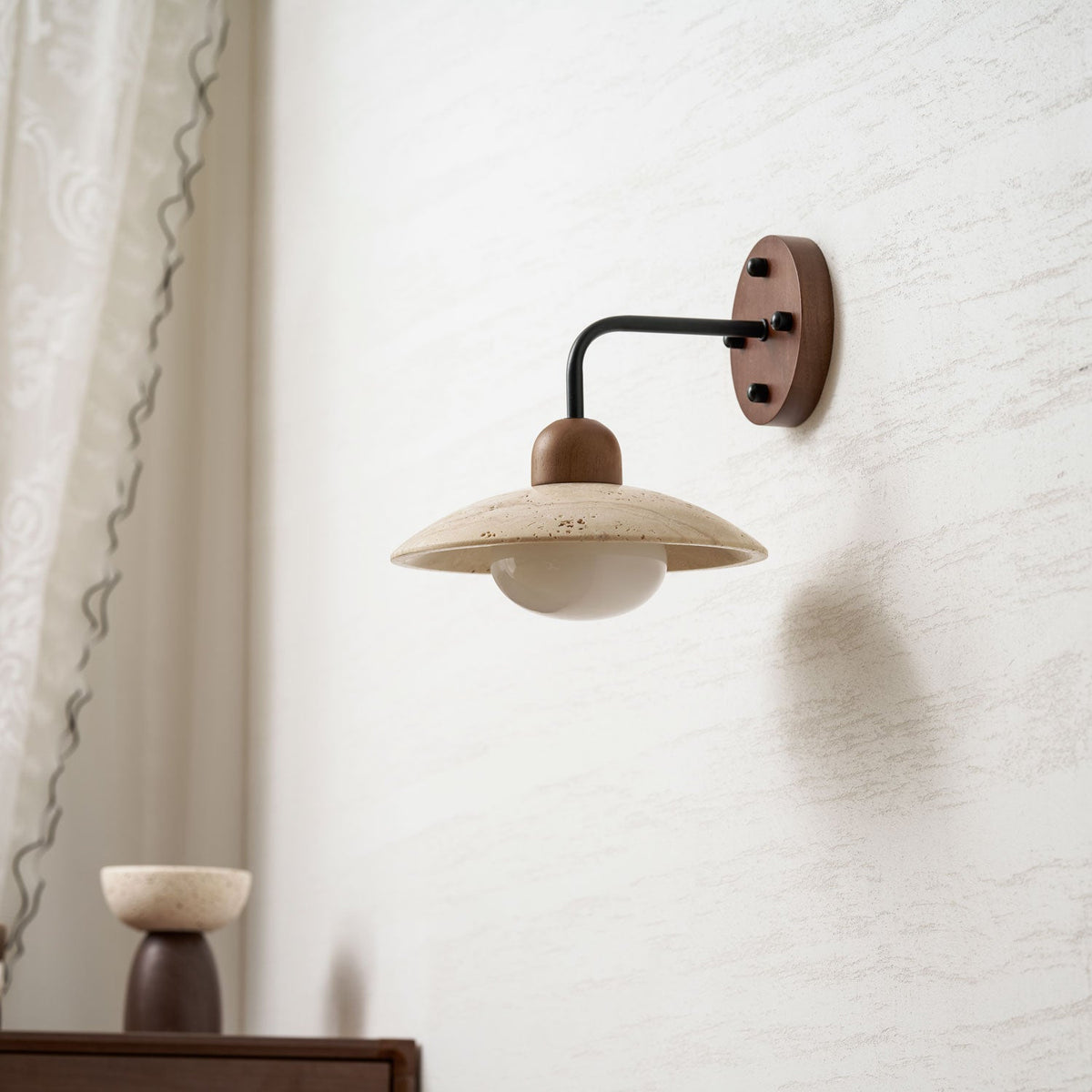 Clérachic wall light with a black arm and a wooden wall plate, its travertine dome unlit against a textured pale wall.