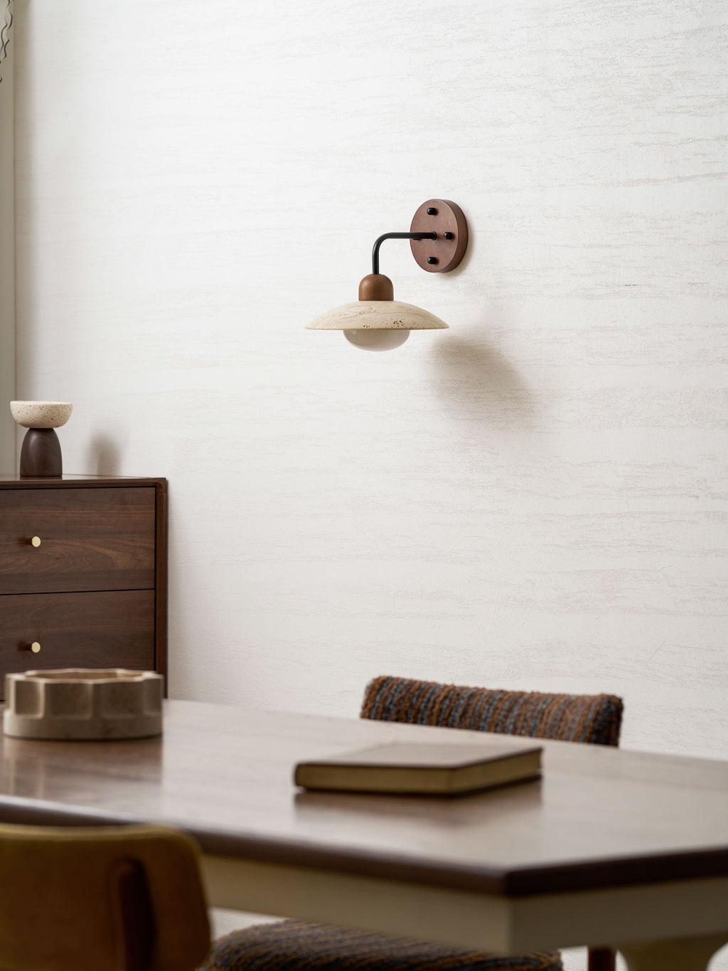 Clérachic travertine wall light mounted high on a textured wall, with a walnut dresser, a stone dish and a table below.
