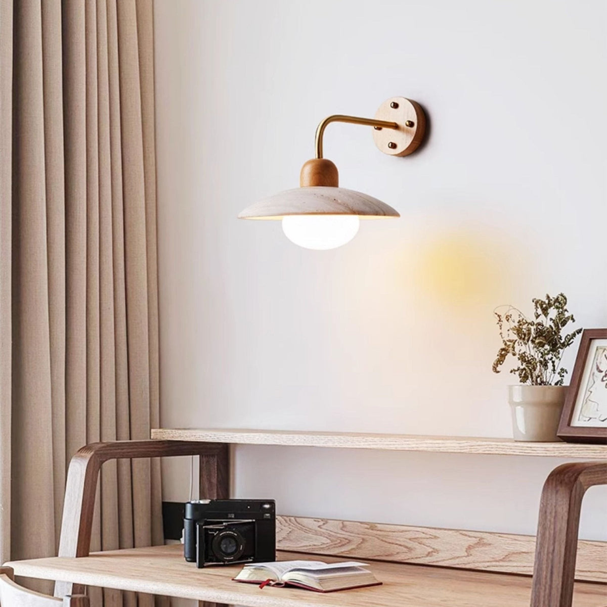 Lit Clérachic wall light with a gold-toned arm above a wooden desk, with a camera, an open book and a potted sprig beside it.