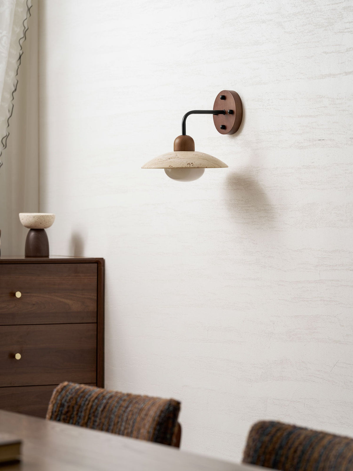 Clérachic wall light with a black arm on a pale textured wall, seen above a walnut drawer unit and woven chair backs.