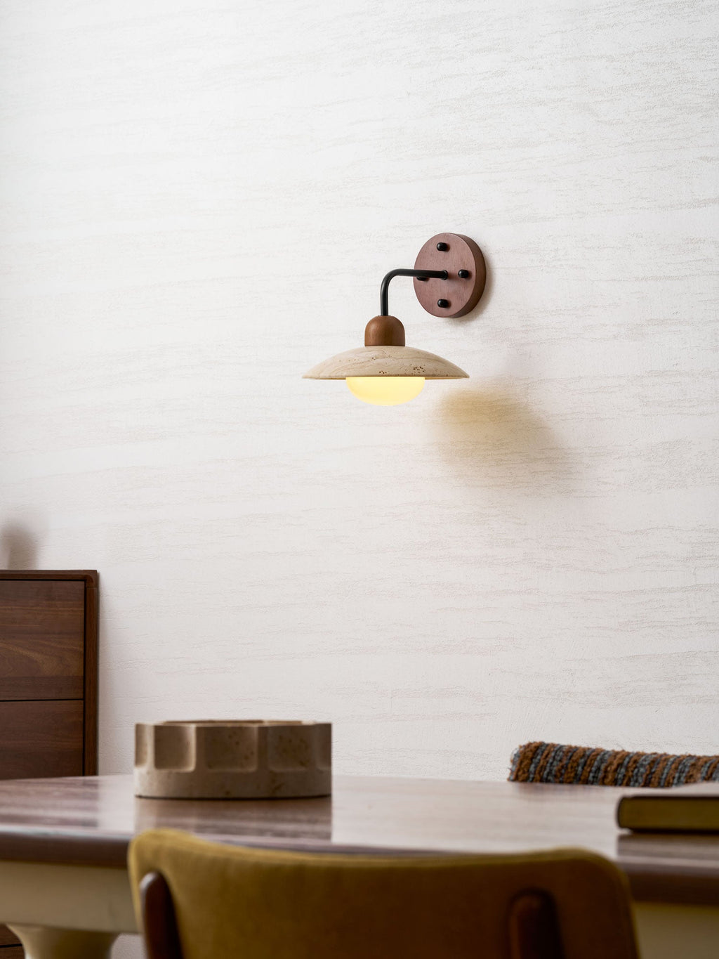 Clérachic wall light glowing softly over a walnut table, with a stone dish and a mustard chair at the bottom of the frame.