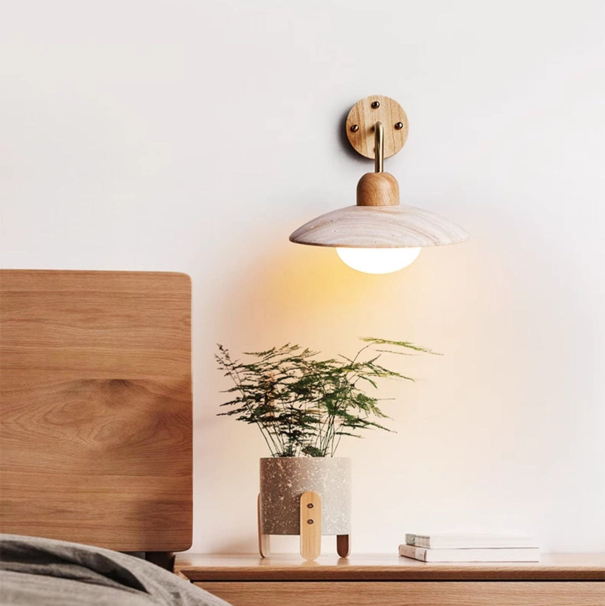 Clérachic wall light glowing above a wooden bedhead and bedside shelf holding a small fern and a stack of books.