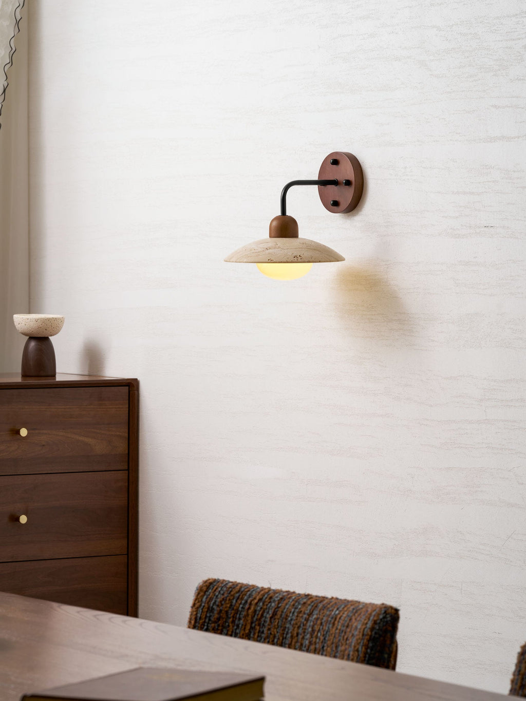 Clérachic wall light on a textured white wall, with a walnut drawer unit and a woven chair in the lower part of the frame.