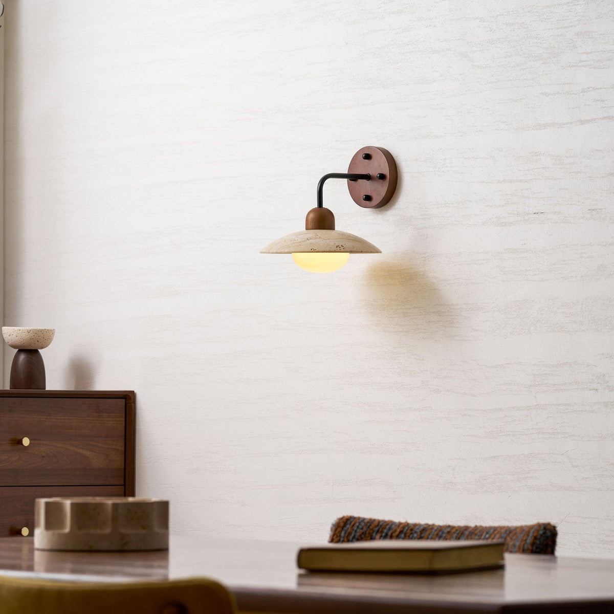 Lit Clérachic wall light with a black arm, mounted over a dining table with a stone dish and a closed book.