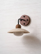 Clérachic wall light with a black arm and a dark walnut plate, the unlit travertine dome casting a soft shadow on the wall.