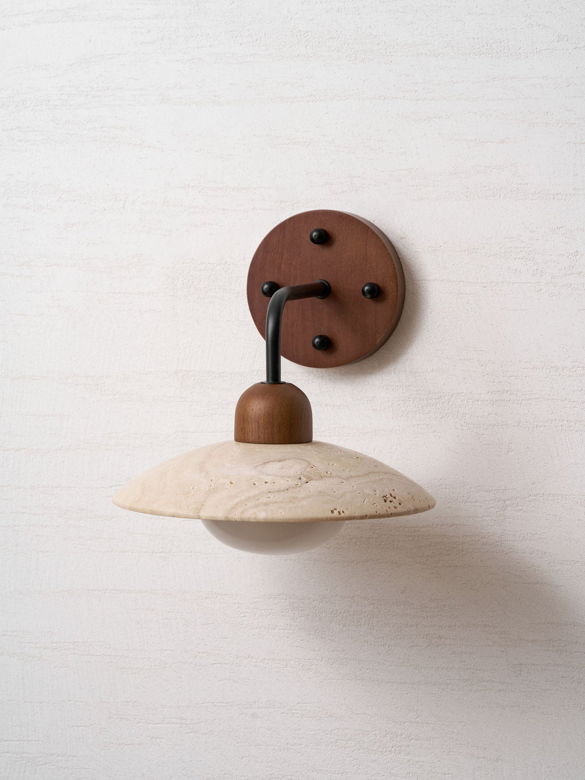 Front view of this Clérachic wall light, with the black arm dropping from a walnut plate to a travertine dome.