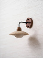 Clérachic wall light seen from slightly below, its travertine dome and white glass bulb unlit on a pale wall.