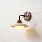 Lit Clérachic wall light with a black arm and a walnut plate, a warm yellow glow under the travertine dome.