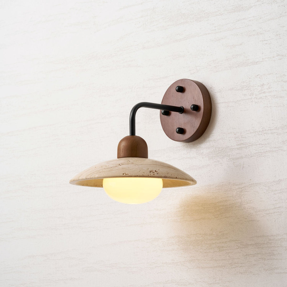 Lit Clérachic wall light with a black arm and a walnut plate, a warm yellow glow under the travertine dome.
