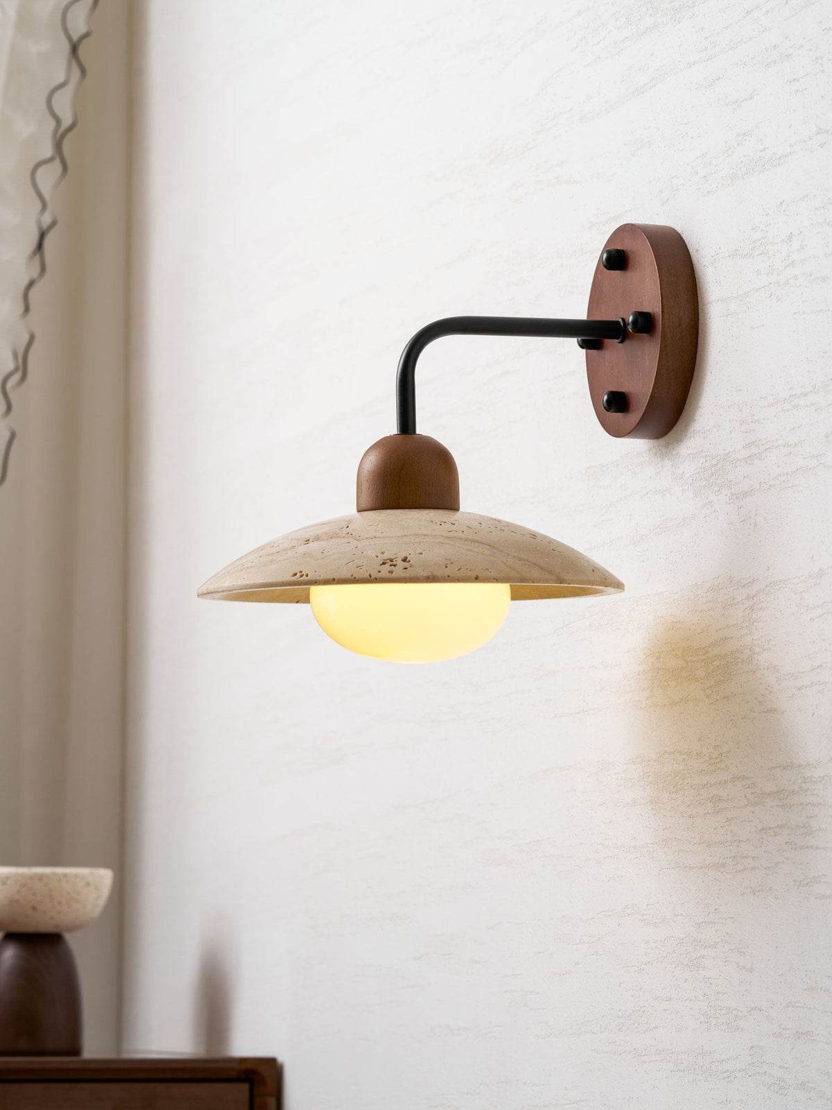 Side view of this Clérachic wall light with a black arm, glowing under its travertine dome beside a curtain edge.