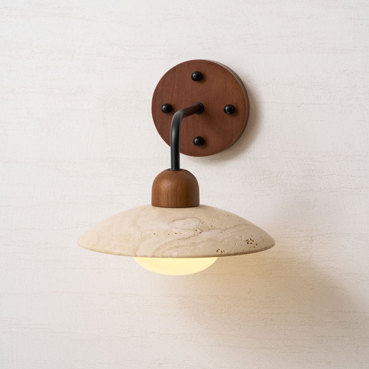 Clérachic wall light with a round walnut plate and four black screws, the travertine dome glowing faintly beneath.