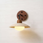 Clérachic wall light with a black arm and a walnut plate, its glass bulb lit warmly under the veined travertine dome.