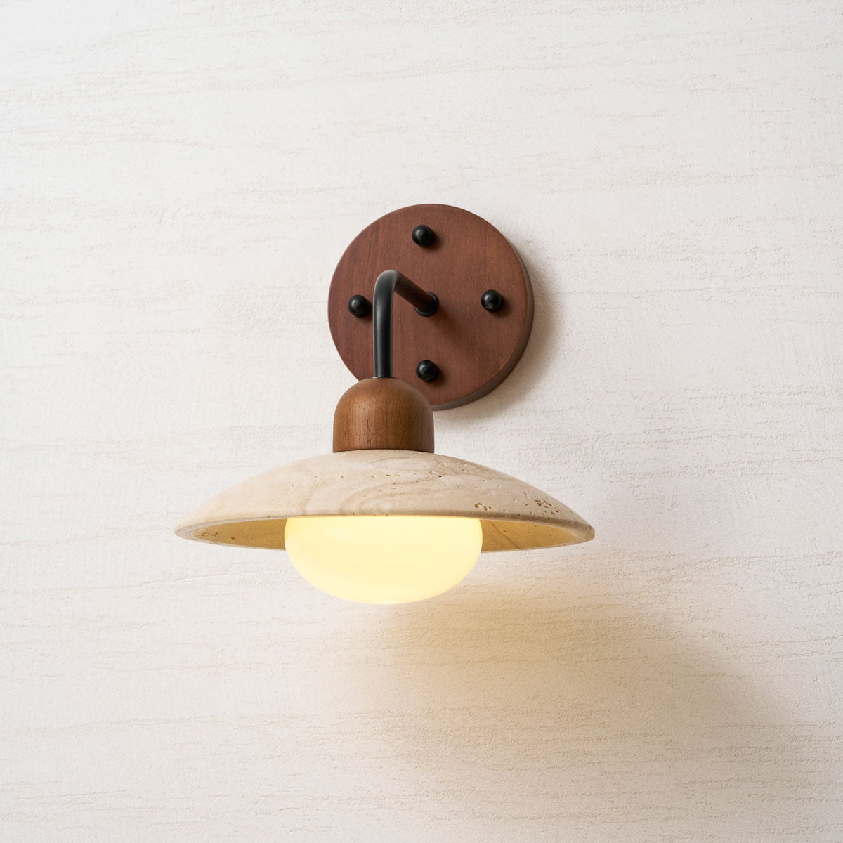 Clérachic wall light with a black arm and a walnut plate, its glass bulb lit warmly under the veined travertine dome.