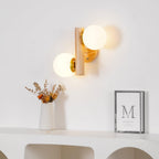 A Clérachic wall sconce with both globes lit, above a white shelf holding a vase of dried flowers and a small framed print.