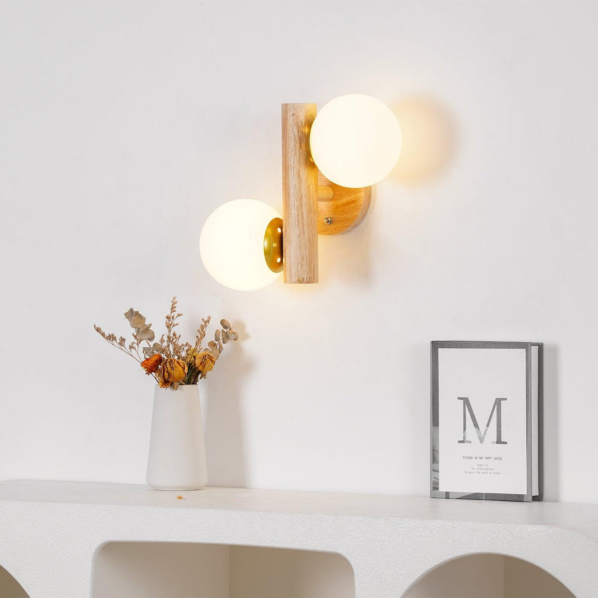 A Clérachic wall sconce with both globes lit, above a white shelf holding a vase of dried flowers and a small framed print.