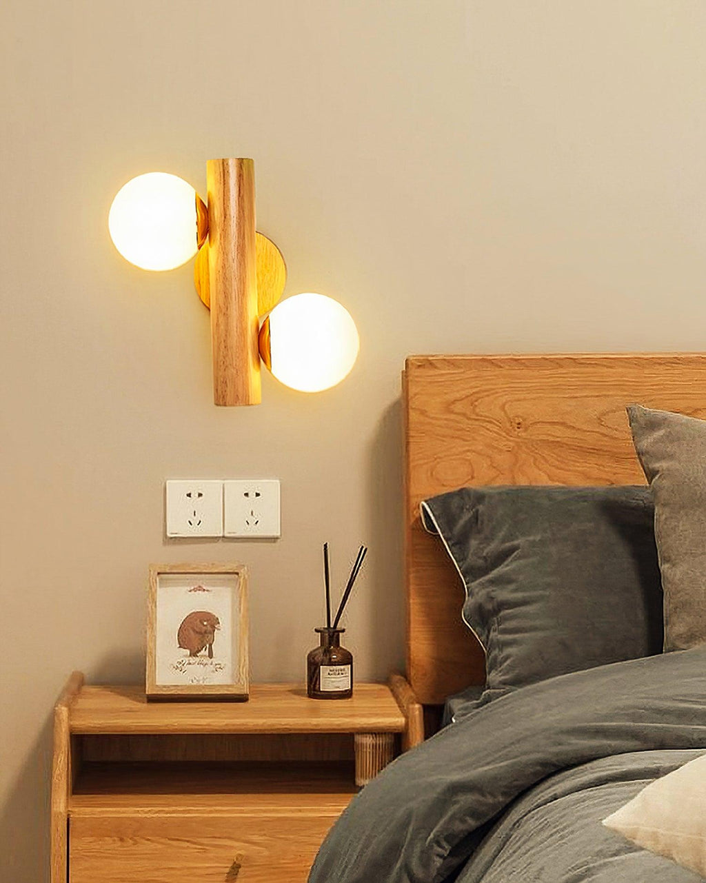 Wood and glass Clérachic wall sconce lit above a wooden nightstand, beside a bed with dark grey linen in a warm beige room.