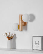 Unlit Clérachic wall sconce with matt white globes beside a vase of dried stems and a framed print on a pale grey wall.