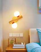 Clérachic sconce with its wooden rod tilted at an angle and both globes lit, above a bedside table with books and a frame.