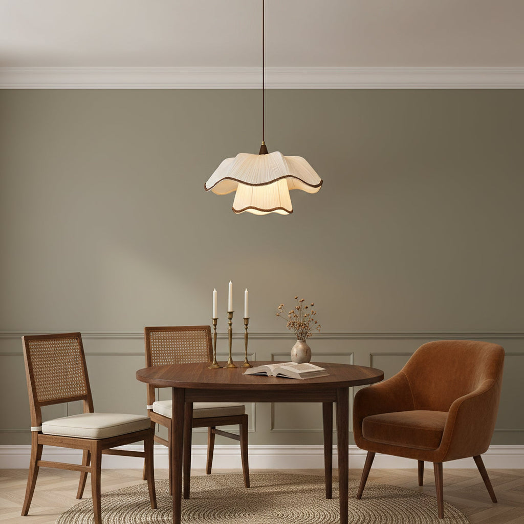 A Clérachic pendant with a ruffled white shade over an oval walnut dining table, with candlesticks, cane chairs and a rust armchair in a sage-green room.