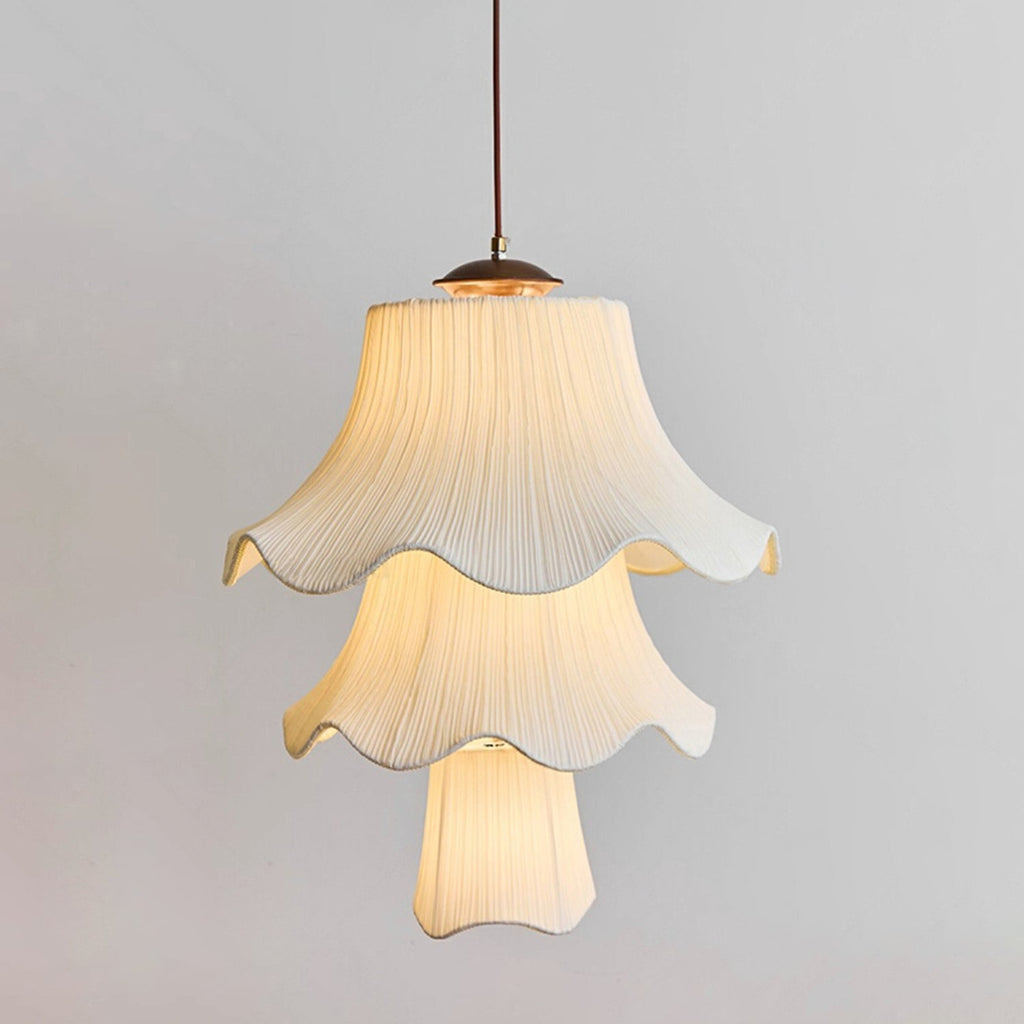 Three tiers of pleated, softly lit fabric shade on this Clérachic pendant, with a wooden canopy and brass fitting against a pale grey wall.