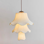 Three tiers of pleated, softly lit fabric shade on this Clérachic pendant, with a wooden canopy and brass fitting against a pale grey wall.