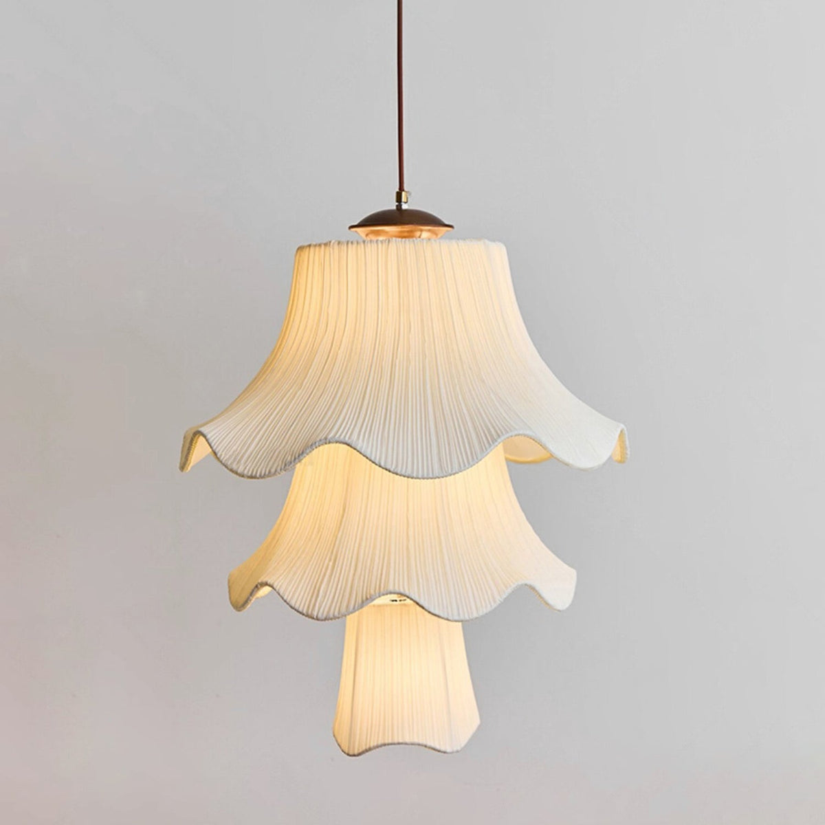 Three tiers of pleated, softly lit fabric shade on this Clérachic pendant, with a wooden canopy and brass fitting against a pale grey wall.