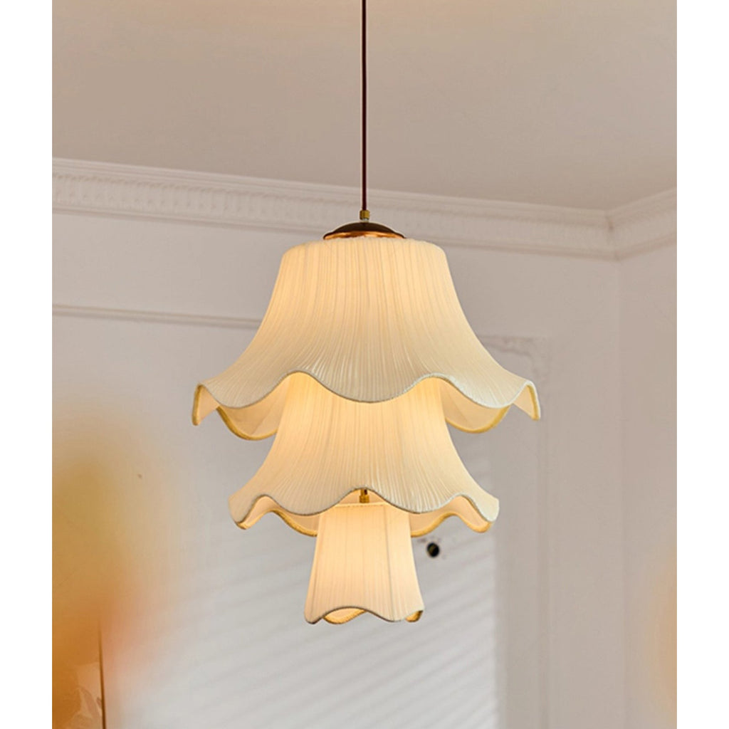 Lit three-tier pleated pendant from Clérachic hanging below a ceiling with white cornice moulding, its scalloped edges glowing warm.