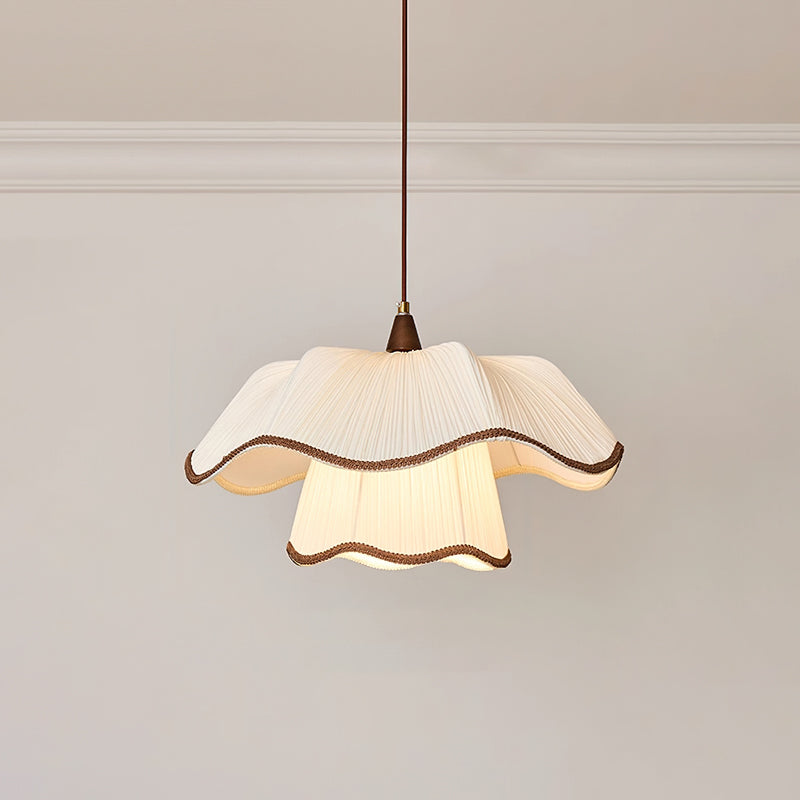 Lit two-tier pleated shade with dark-brown piped edges on a Clérachic pendant, hanging against a pale wall under white moulding.