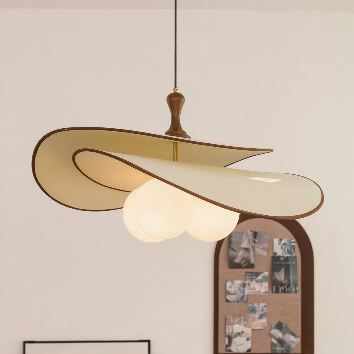Beige version of this Clérachic pendant with a thin brown edge, its three opal globes lit, hanging in front of an arched pinboard.