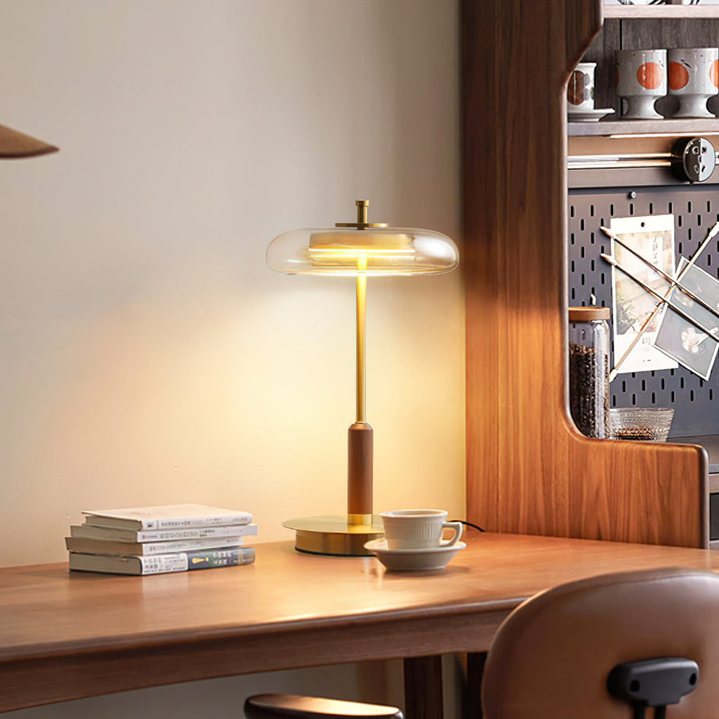 Lit Clérachic table lamp on a walnut desk beside a stack of books and a cup, its glass shade glowing warm against a cream wall.