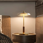 Clérachic table lamp glowing on a round bedside table, the brass base and walnut stem set against a woven headboard.
